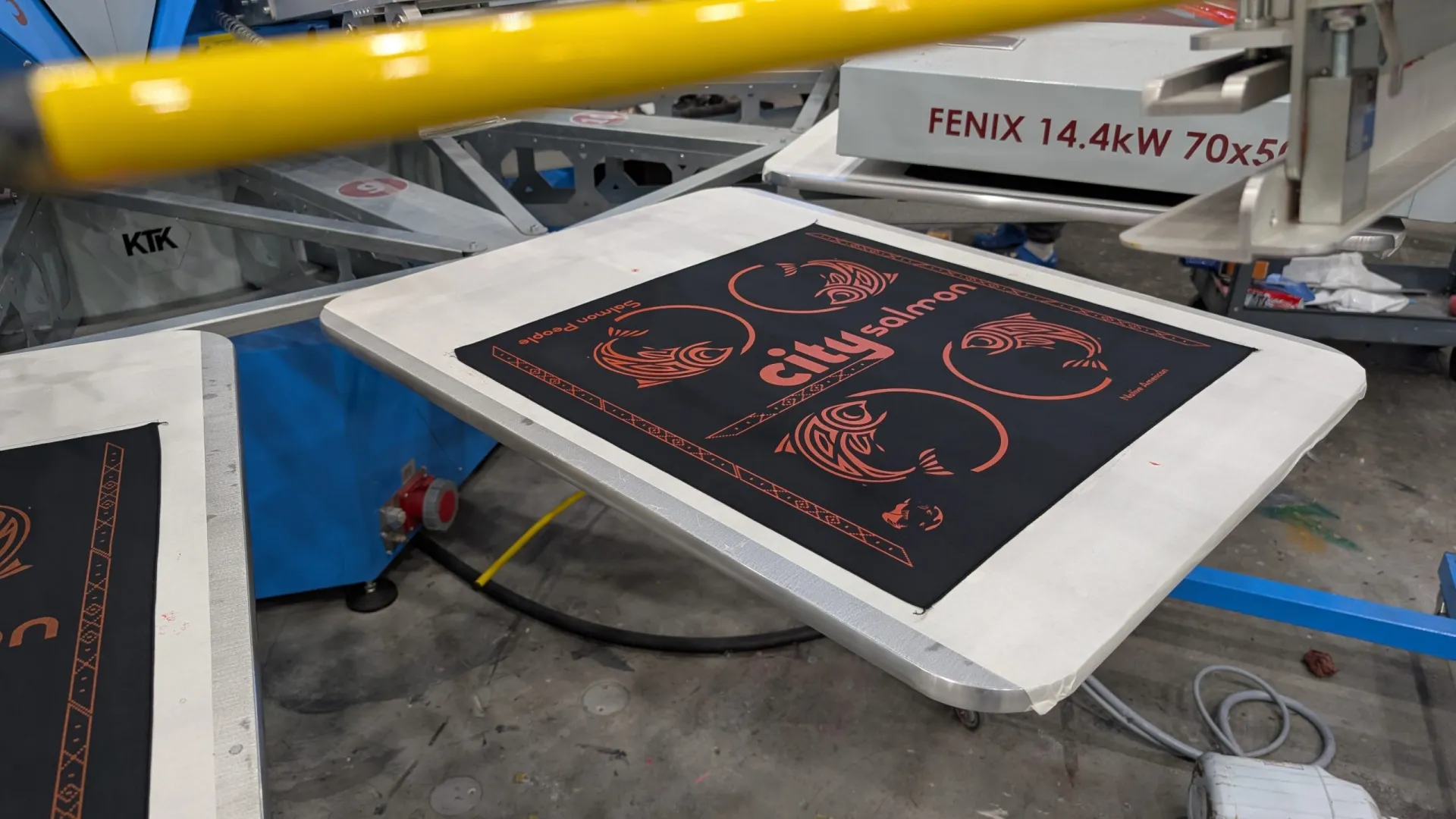 Screen printing press producing black cotton custom bandanas with orange ink at AMBRO Manufacturing for Canadian clients