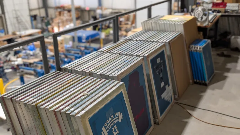 A row of aluminum screen printing frames with mesh screens stacked neatly on a mezzanine inside AMBRO Manufacturing’s New Jersey print shop, overlooking the production floor below.