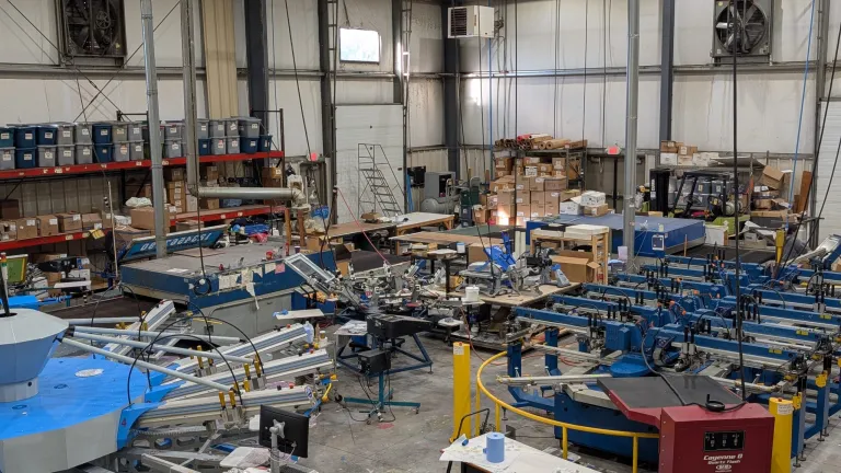 Overhead view of the AMBRO Manufacturing factory showing multiple automatic screen printing presses, conveyor dryers, and organized workstations inside a New Jersey apparel production facility.