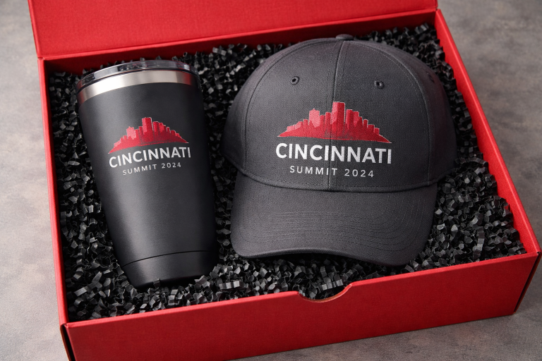Red swag box with engraved tumbler and embroidered baseball cap on black shredded paper
