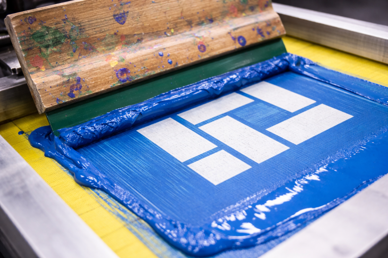 Closeup of a screen printing screen with blue ink and a squeegee spreading ink across the mesh stencil.