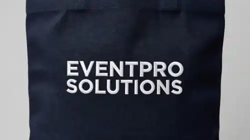 A close-up view displays a high-quality navy canvas tote bag featuring a crisp white embroidered company logo. The bright thread stands out sharply against the deep fabric under professional studio lighting.